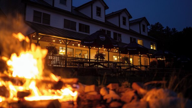 A building with a covered outdoor seating area and a fire in the foreground. - Inn By The River (West Forks)