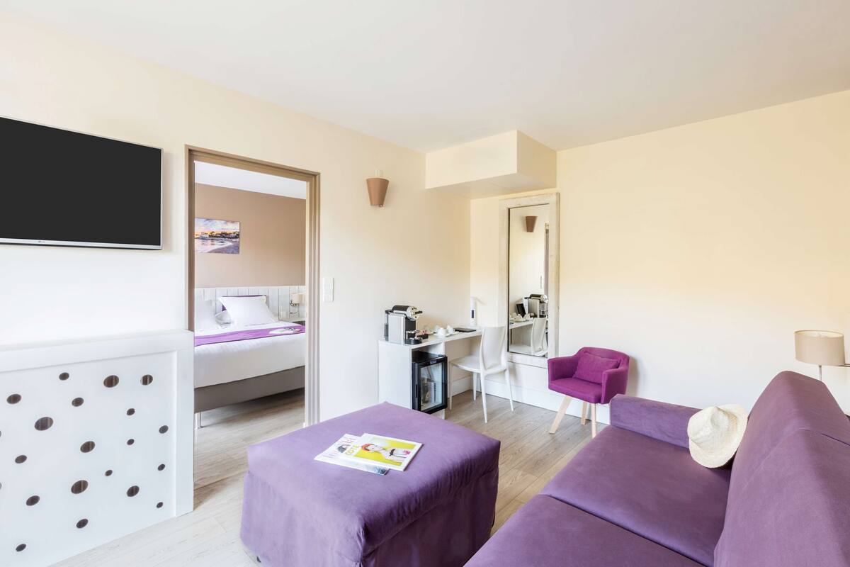 A modern hotel room with a bed, a desk, a purple sofa, and a small table with a magazine.