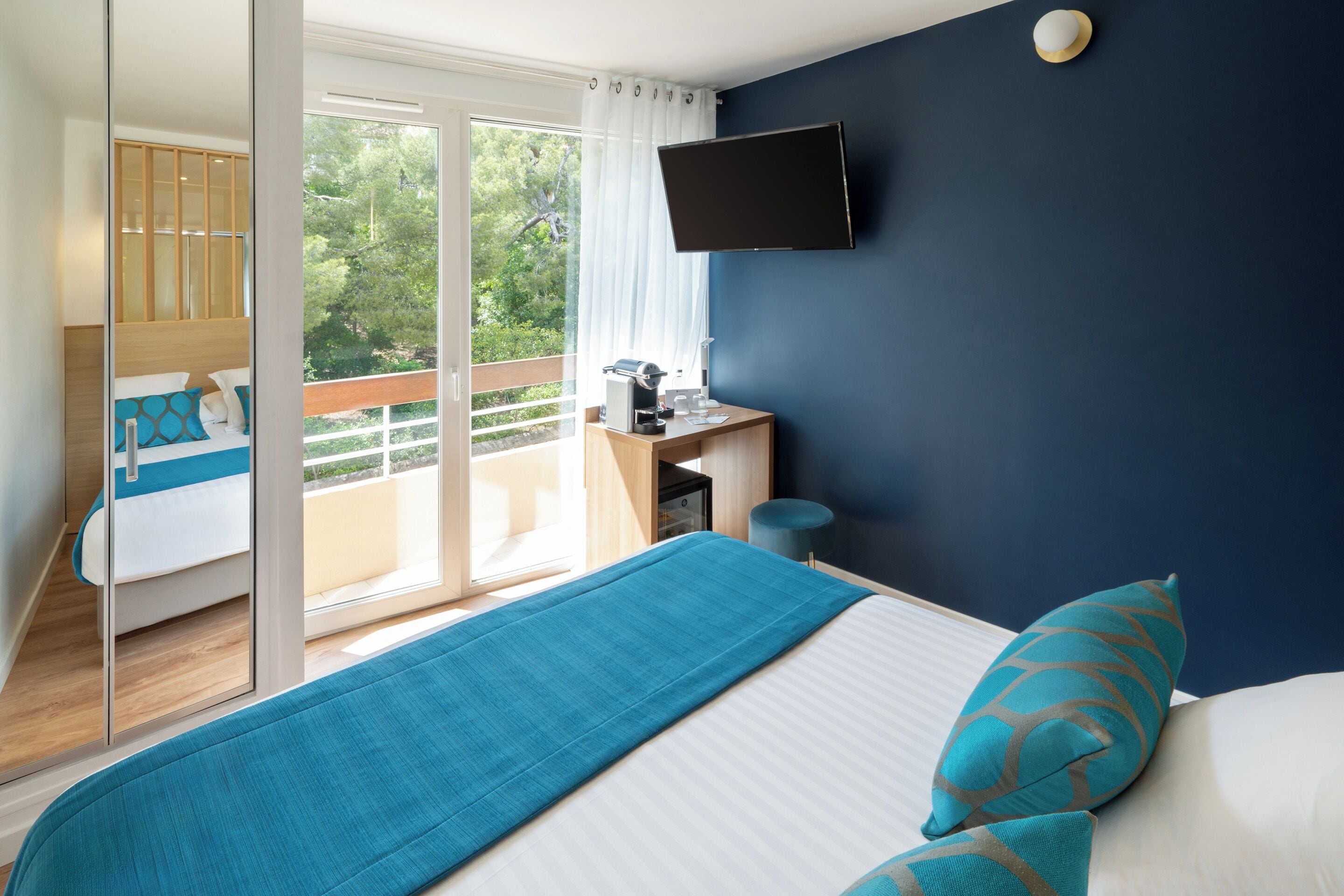 A bedroom with a bed, a desk, a chair, a TV, and a balcony with a view of trees.