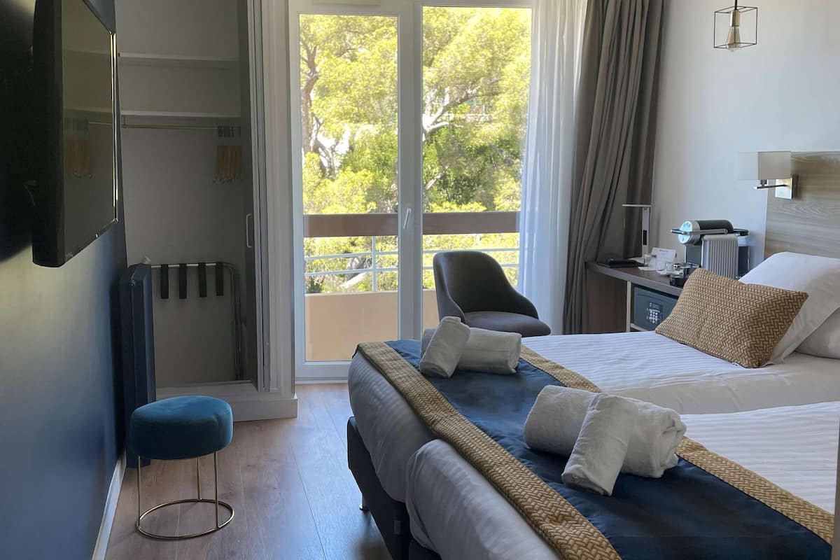 A hotel room with a large bed, a desk with a chair, a TV mounted on the wall, and a view of trees through the window.