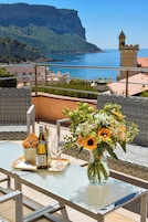 Una mesa con una botella de vino, una copa y un ramo de flores en un balcón con vista al mar y edificios, incluyendo una torre.