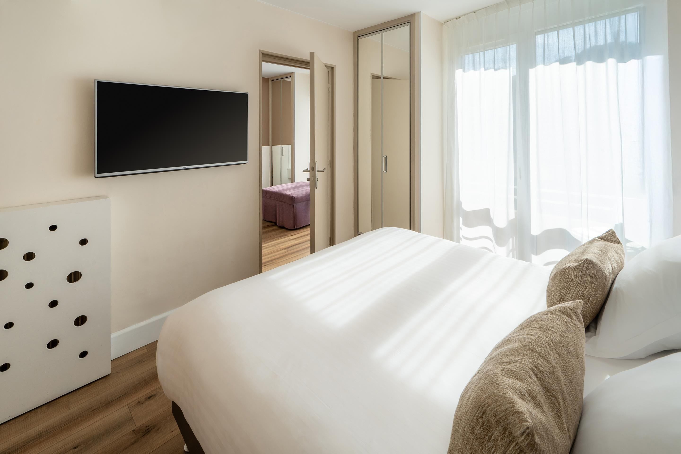 A modern hotel room with a large bed, a flat-screen TV mounted on the wall, and a view of the hallway.
