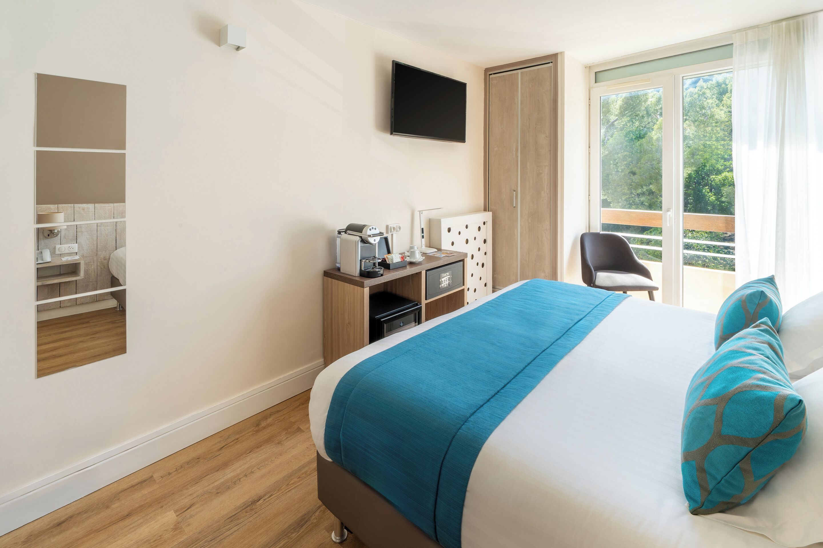 Ein Hotelzimmer mit Bett, Schreibtisch mit Computer, Stuhl, Fernseher und einem Balkon mit Blick auf Bäume.