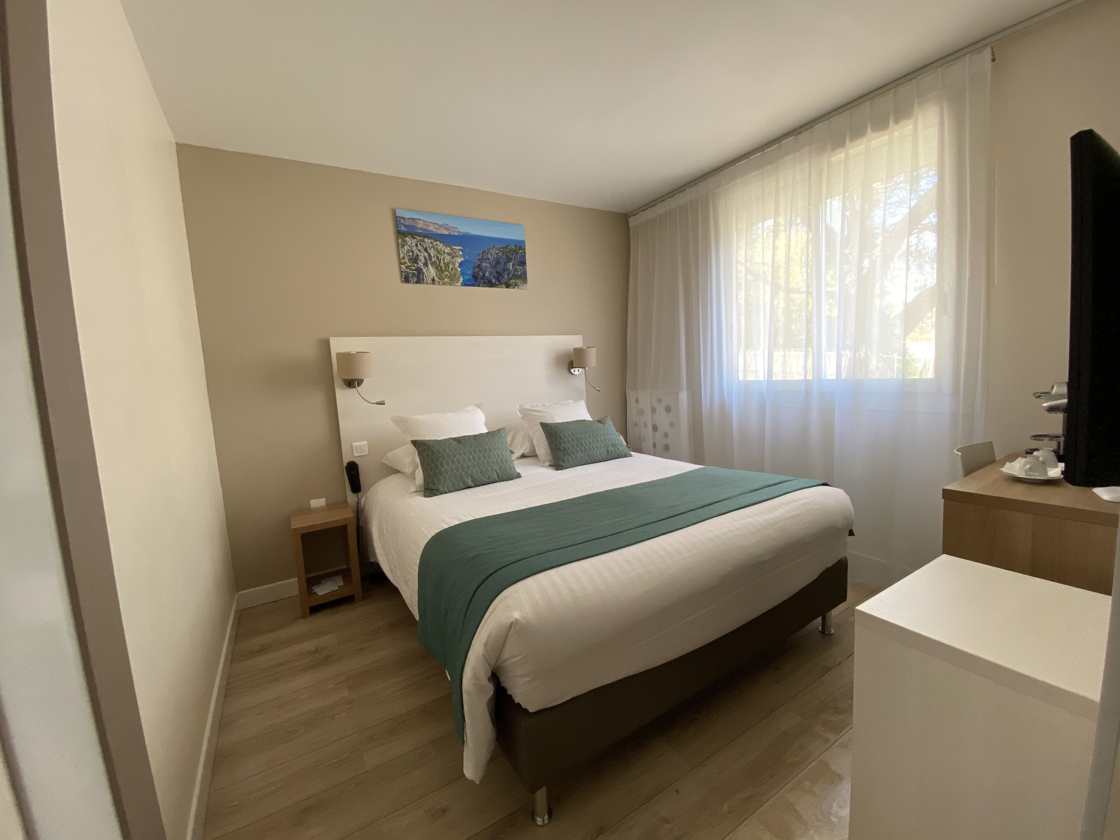 Habitación de hotel ordenada con una cama grande, una mesita de noche, una mesita auxiliar pequeña, un escritorio con una silla, una ventana con cortinas translúcidas y un cuadro en la pared.