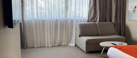 A modern hotel room with a sofa, a bed, a small round table, and a red ottoman.