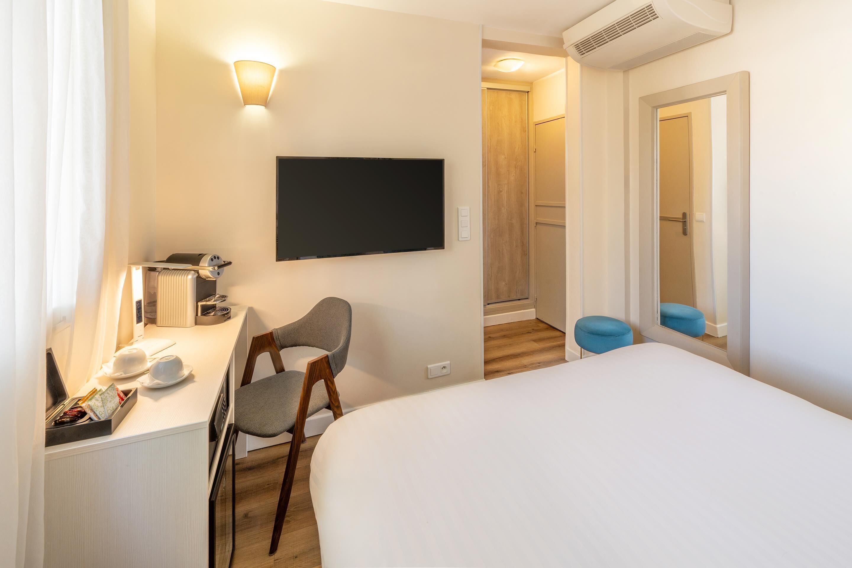 A hotel room with a bed, a desk with a chair, a flat-screen TV, a radiator, and a small stool.