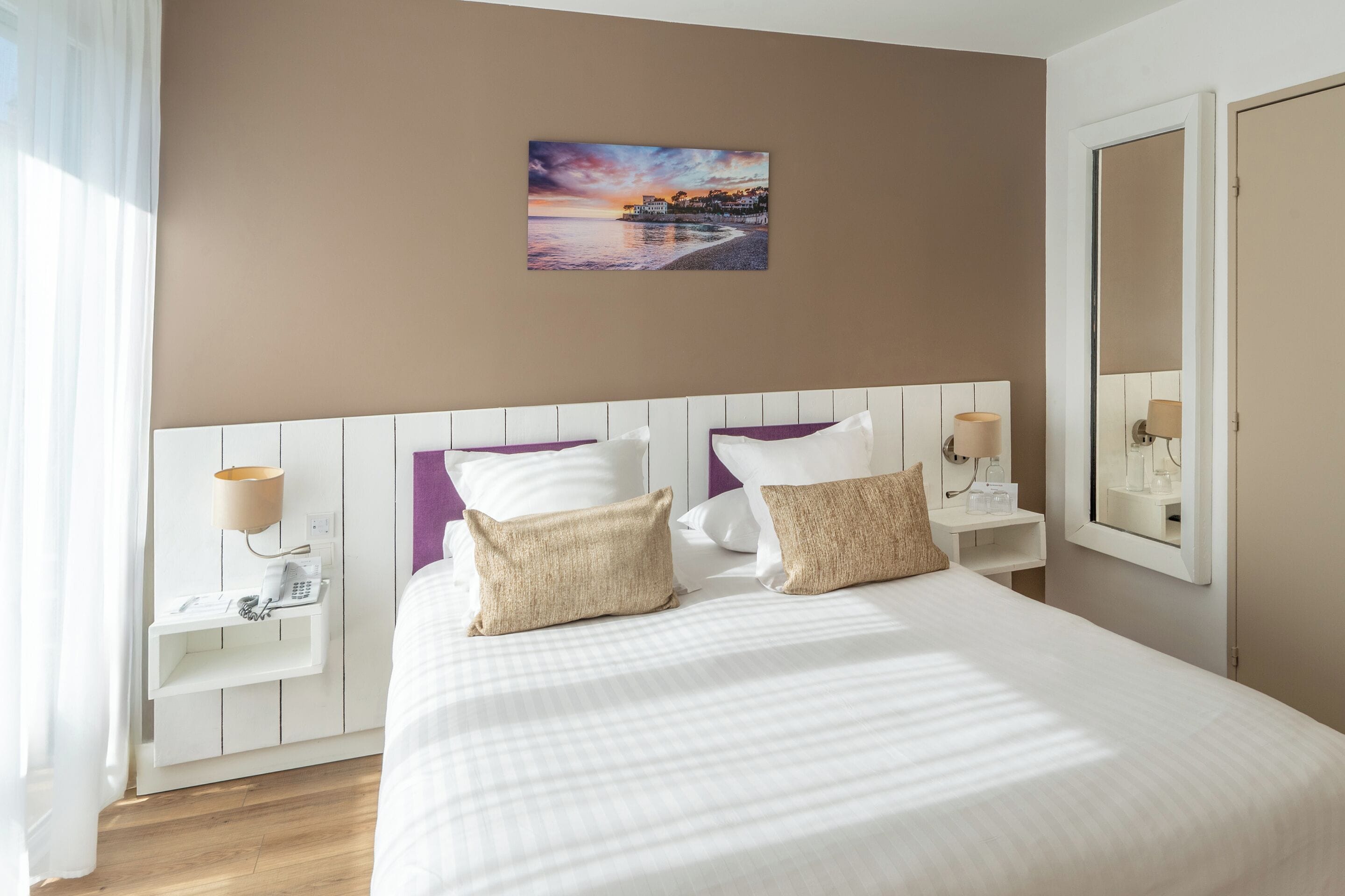 A neatly arranged hotel room with a large bed, two bedside tables, a nightstand, a wall-mounted lamp, a mirror, and a painting of a sunset over a beach.