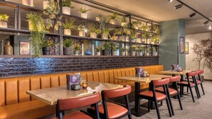 A restaurant interior with a brick wall, wooden tables, red chairs, and shelves with potted plants.