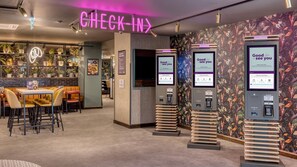 A check-in area with a neon sign, seating, a menu board, and self-service kiosks.