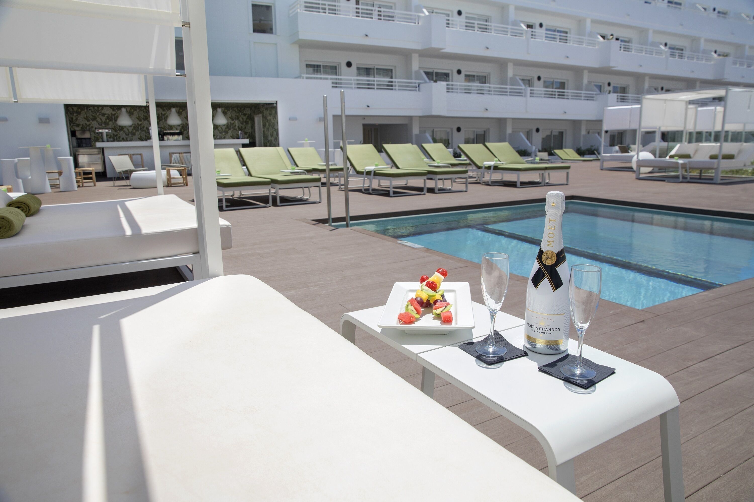 A poolside area with lounge chairs, a table with a bottle of champagne and glasses, and a plate of fruit.