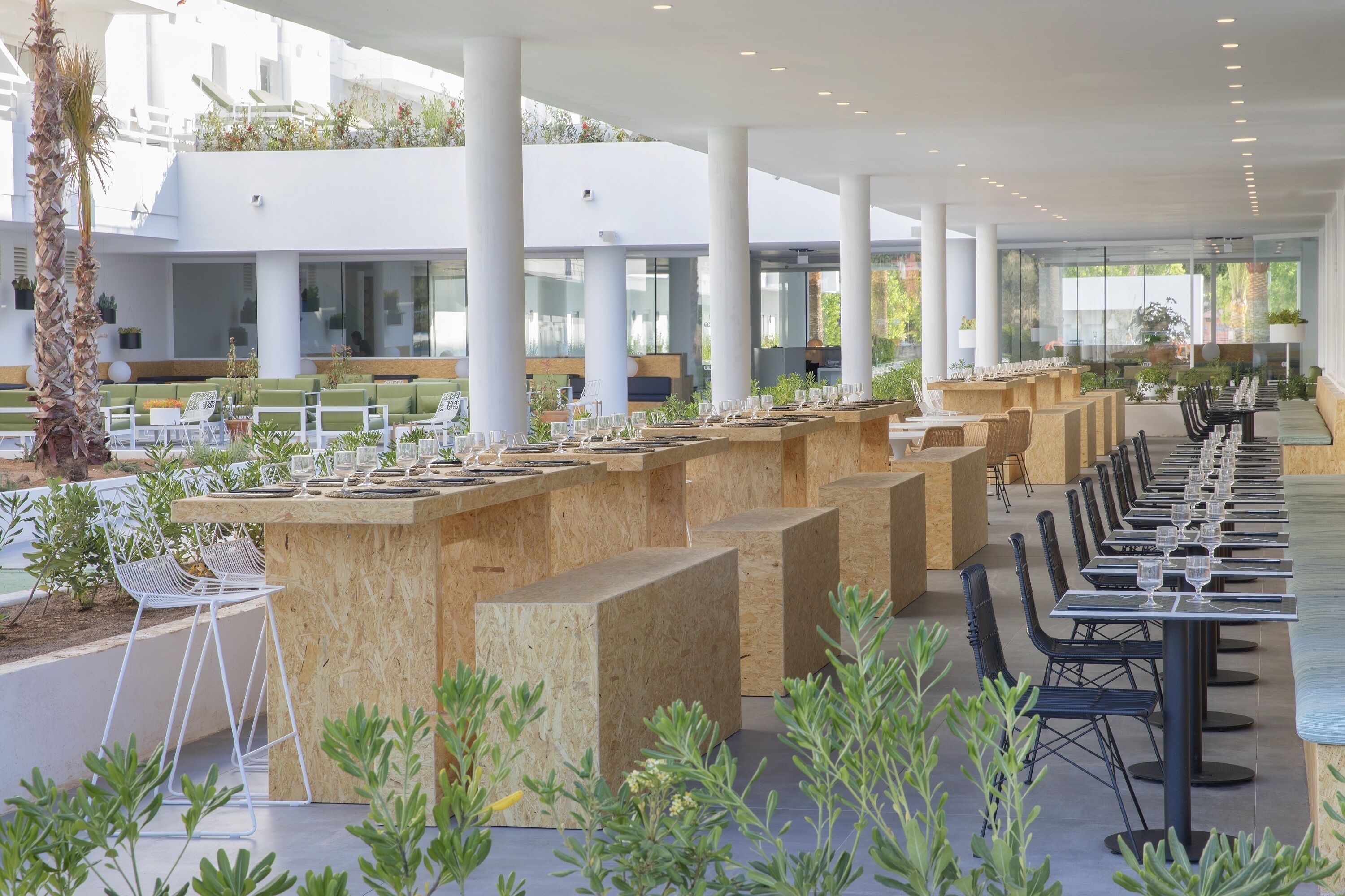 Een moderne buiten eethoek met meerdere houten tafels en stoelen, omgeven door groen en witte pilaren.