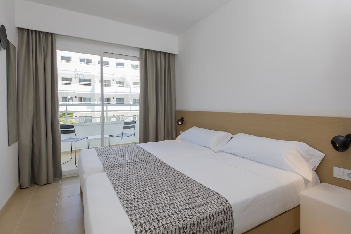 A hotel room with a large bed, a bedside table, a lamp, and a view of a building through the window.