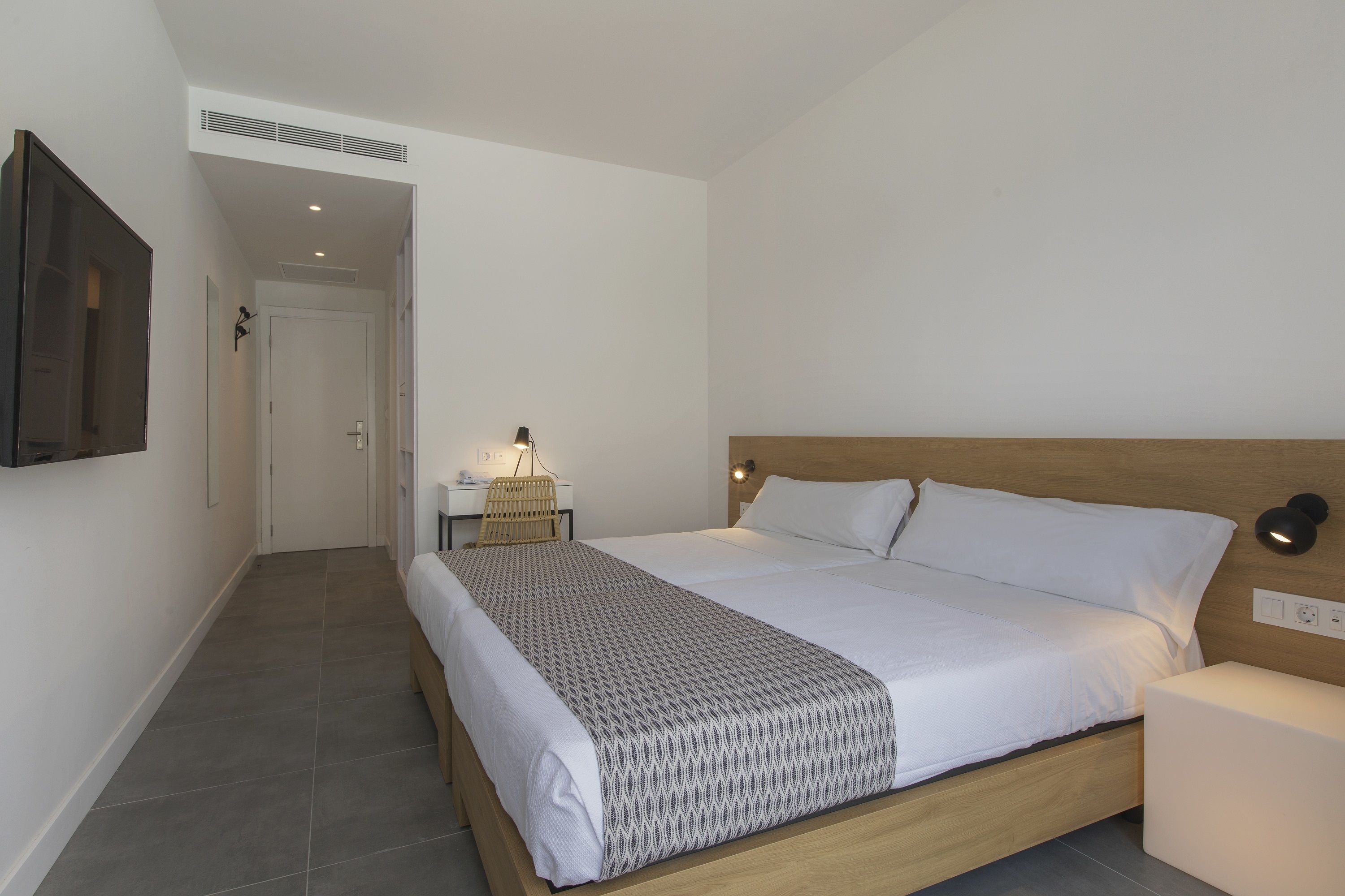A bedroom with a wooden bed, white bedding, a bedside lamp, and a wall-mounted TV.