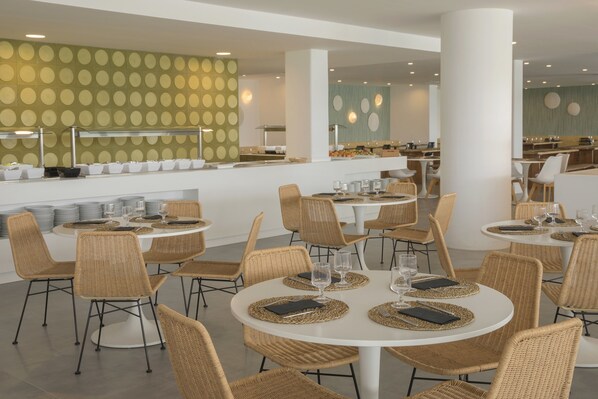 Un comedor moderno con mesas redondas, sillas de mimbre y un mostrador de buffet con platos y tazones.