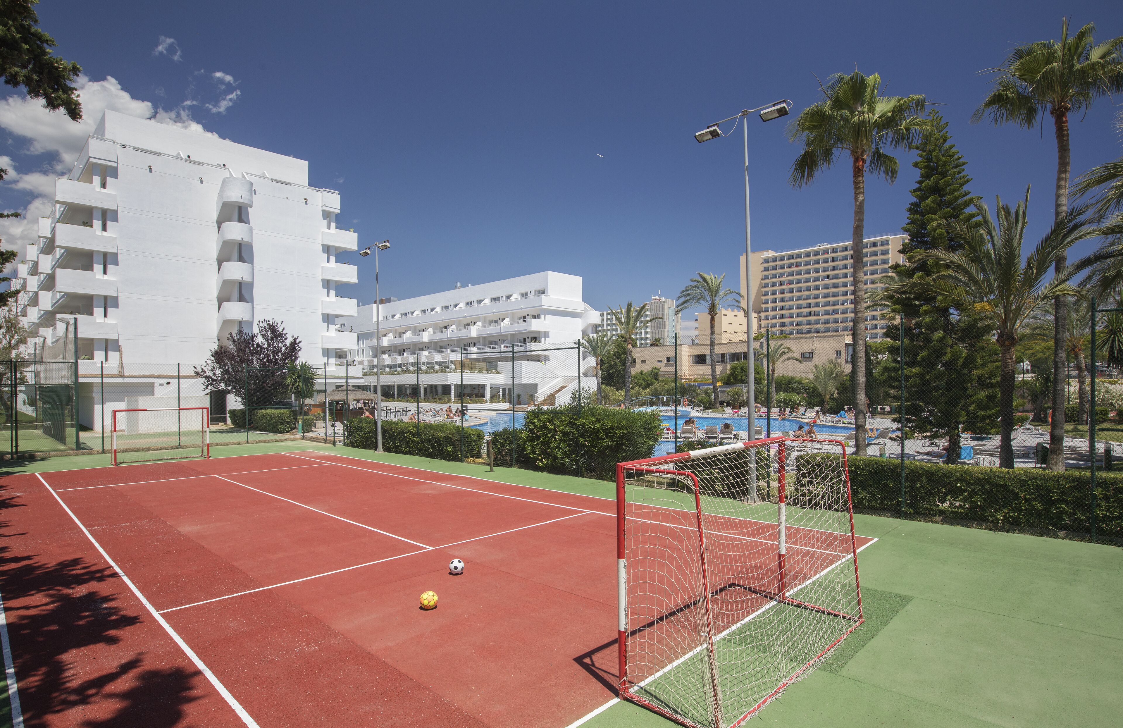 A tennis court with a goal post, surrounded by palm trees and buildings.