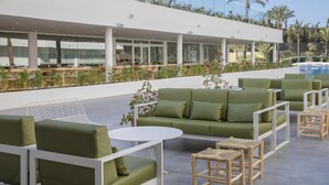 A modern outdoor seating area with green sofas and white tables, overlooking a pool and palm trees. - HM Martinique Aparthotel (Calvia)