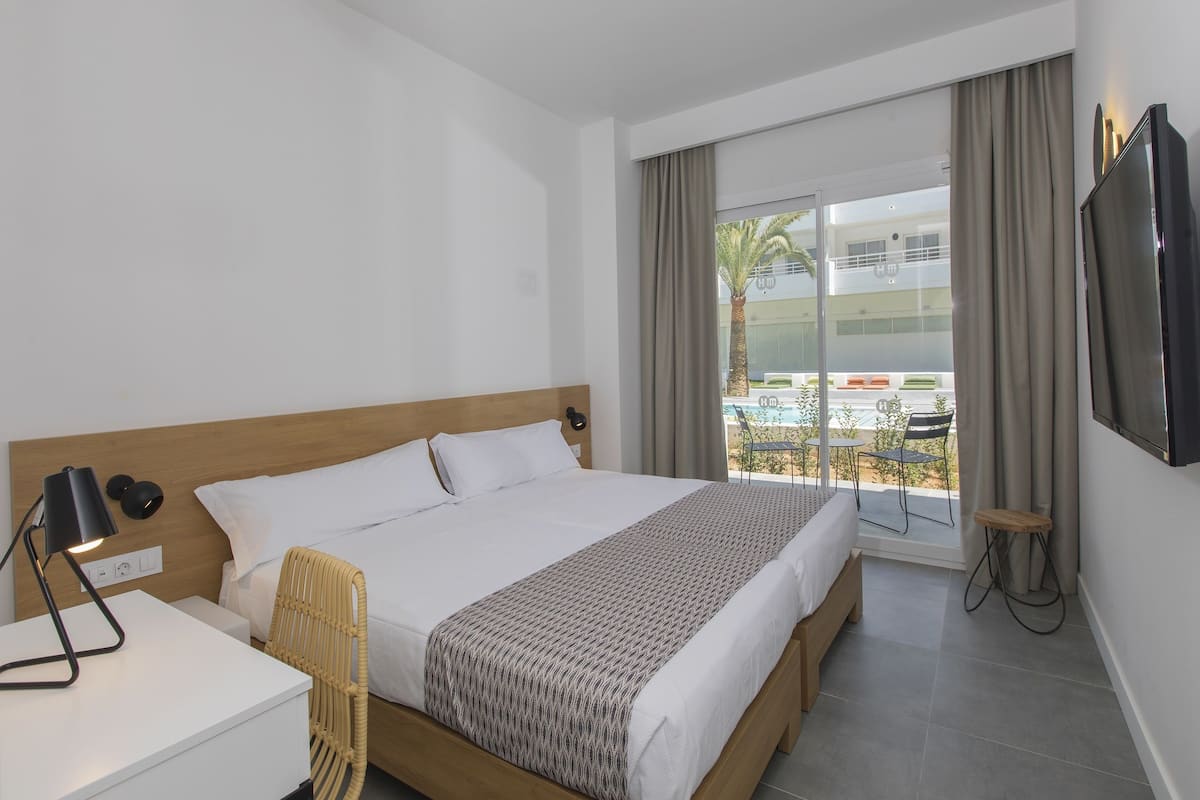 A hotel room with a large bed, a bedside table with a lamp, a TV mounted on the wall, and a balcony with a view of the outside.