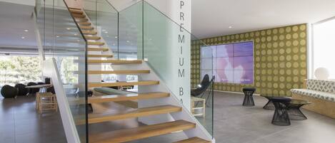 A modern interior with a glass staircase, a large screen displaying a hotel, and a seating area with a patterned wall.