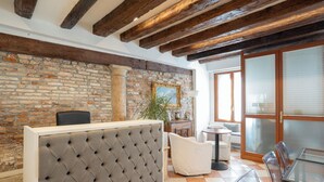 Zona de recepción con pared de ladrillo, vigas de madera, cabecero tapizado, una silla blanca, una mesa de vidrio y una planta.