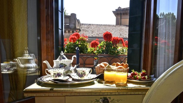Desayuno dispuesto con juego de té, pan y fruta sobre una mesa junto a una ventana con vistas a flores rojas y un edificio.