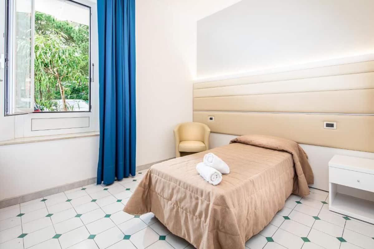Habitación de hotel con cama, una silla, una mesita de noche, ventana con cortinas y vista a zonas verdes.