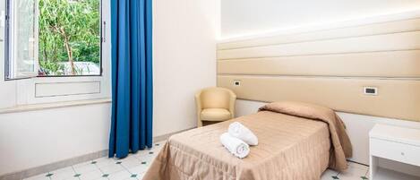 A hotel room with a bed, a chair, a nightstand, a window with curtains, and a view of greenery outside.