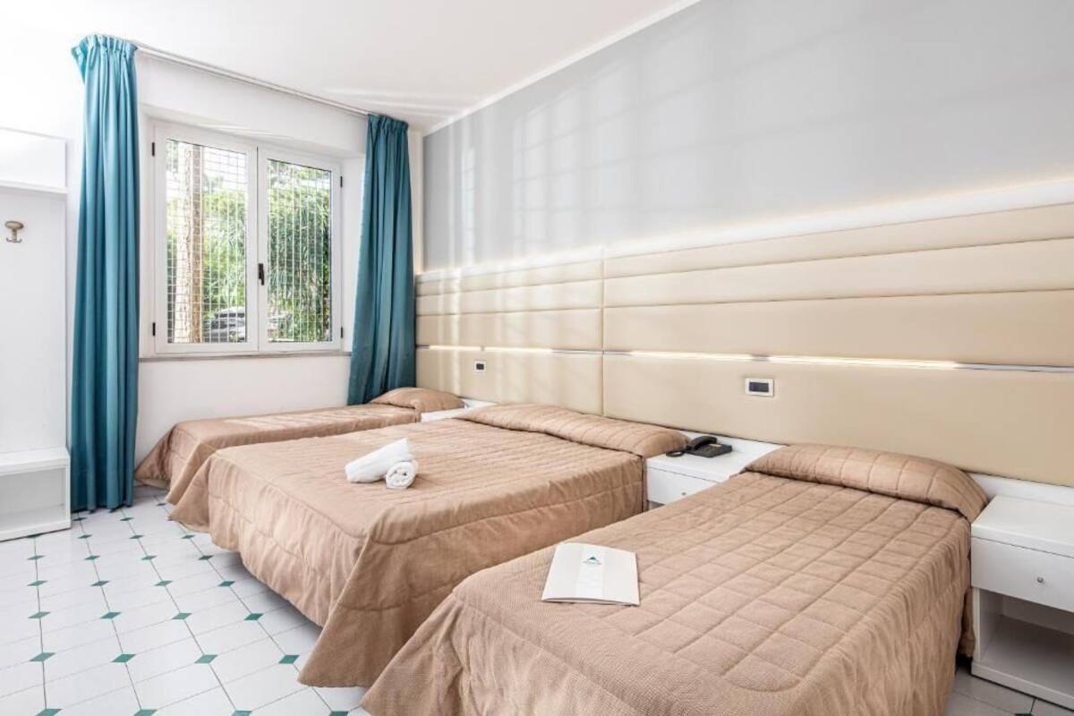 Habitación de hotel con dos camas, una ventana con cortinas, una mesita de noche y una pared blanca con un panel de luz.