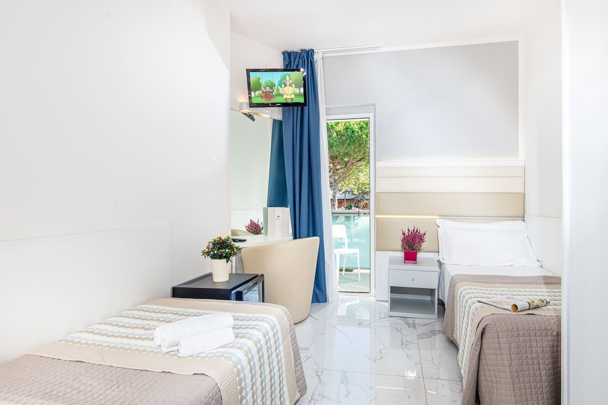 Una habitación de hotel moderna con dos camas, un televisor montado en la pared, un pequeño escritorio con una planta y un balcón con vistas a zonas verdes.