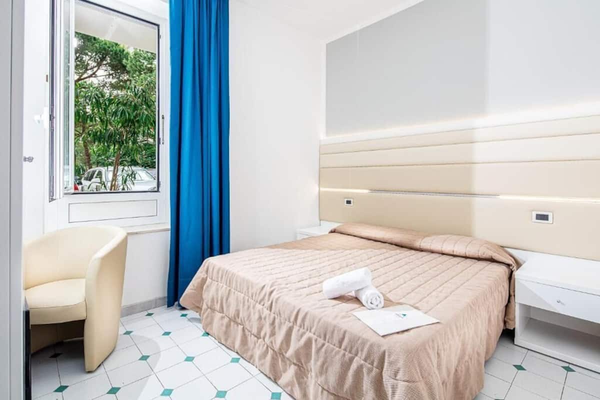 Una habitación de hotel moderna con una cama, una silla, un ventanal con cortinas azules y vistas a árboles.
