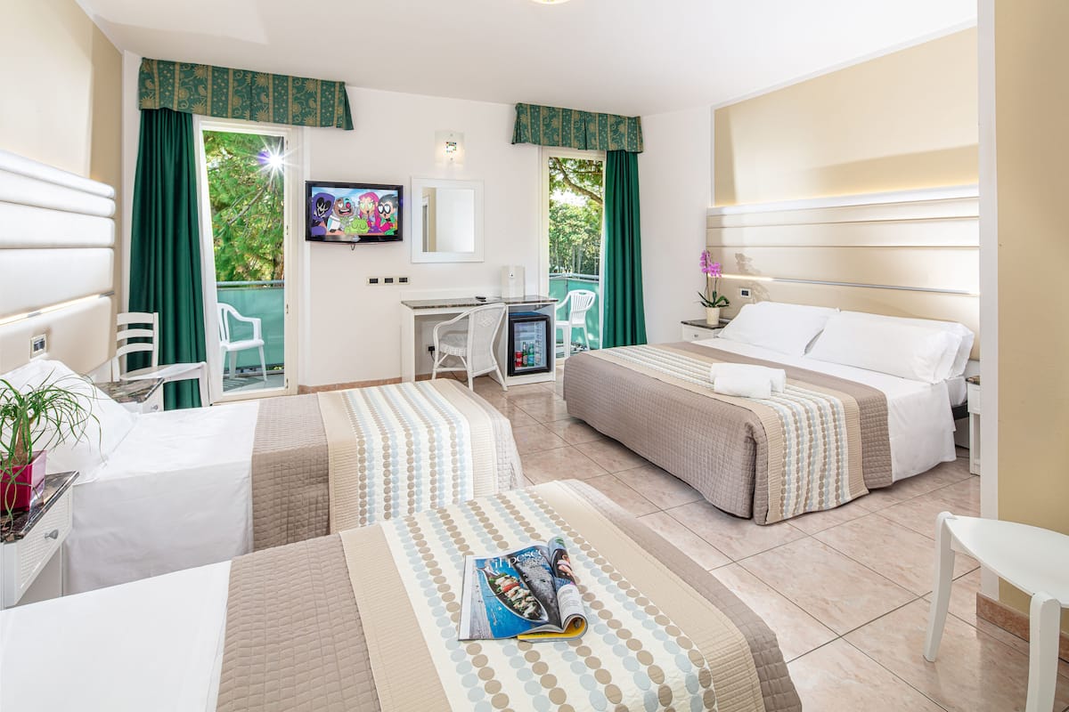 Habitación de hotel con dos camas, un escritorio, un televisor y un balcón con cortinas verdes.