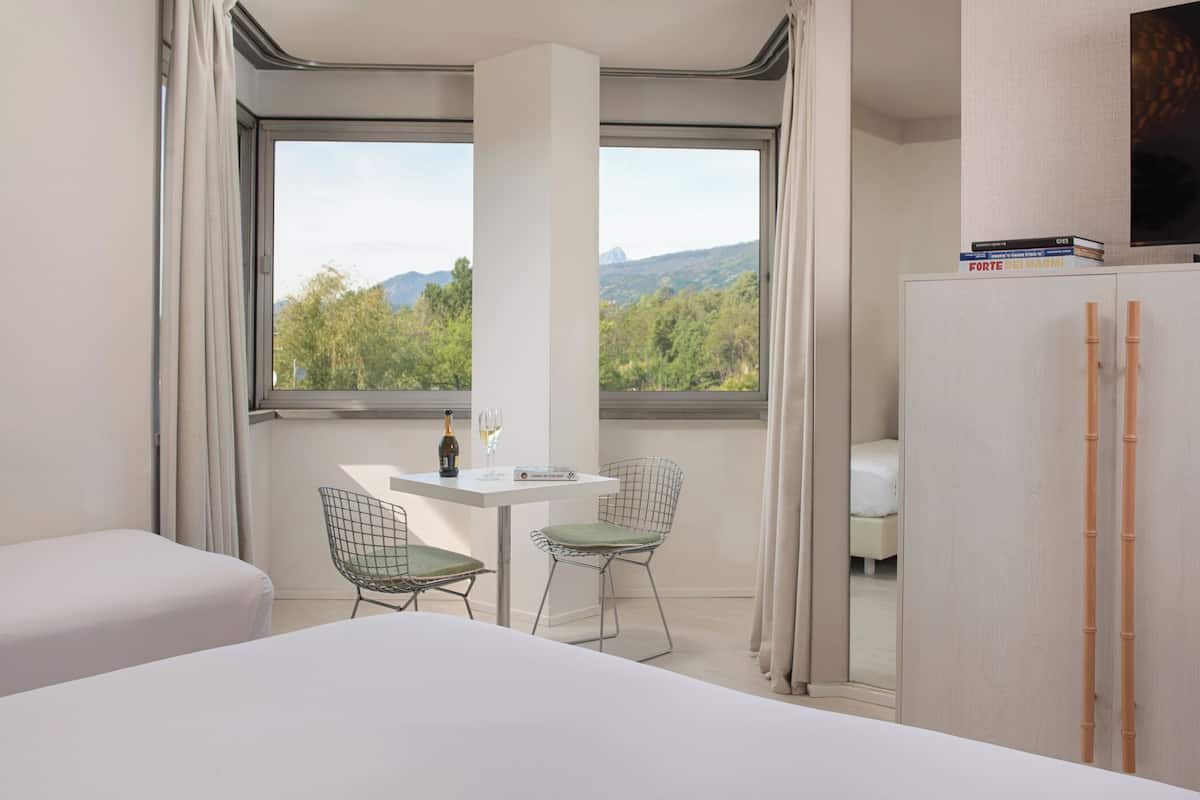 A hotel room with a bed, a small table with two chairs, a minibar, a TV, and a window with a mountain view.