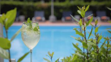 A glass of a light-colored beverage with mint leaves, placed near a poolside with greenery and lounge chairs in the background.