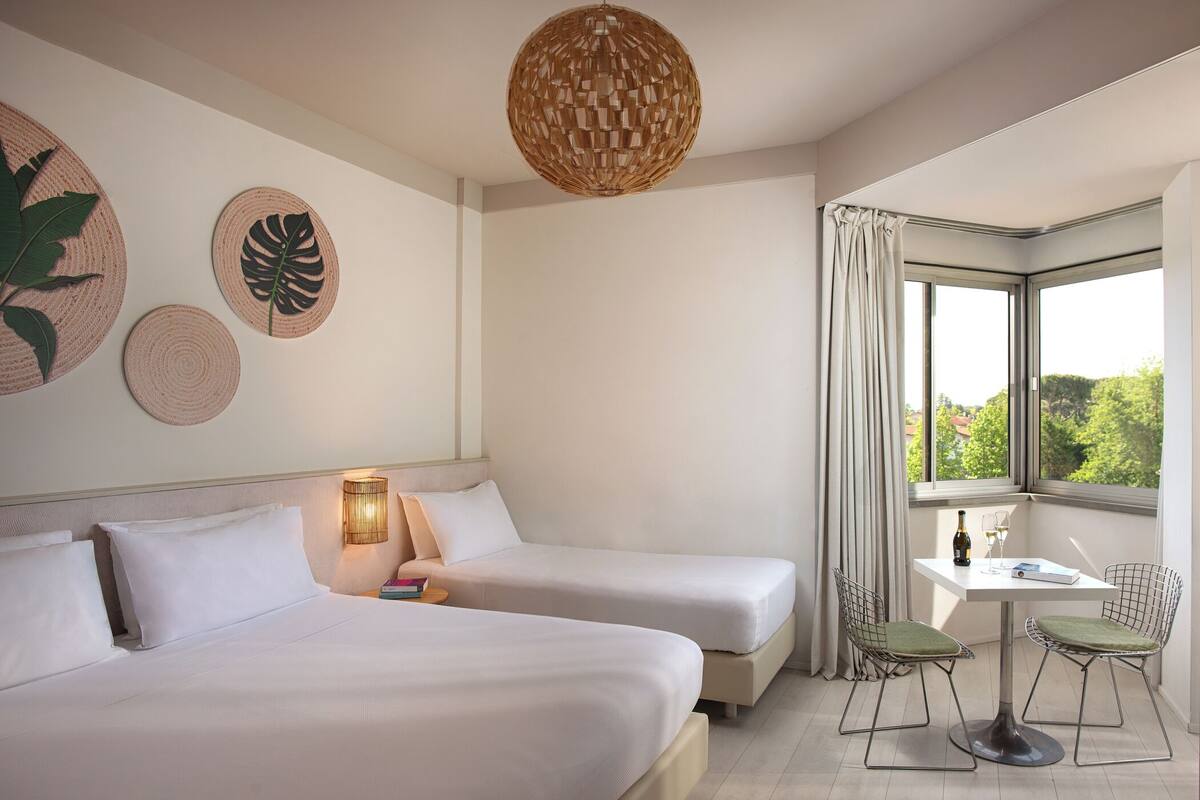 A hotel room with two beds, a small table with chairs, a bottle of wine, and a view of greenery through a window.