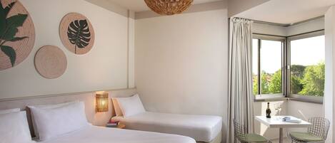 A hotel room with two beds, a small table with chairs, a bottle of wine, and a view of greenery through a window.