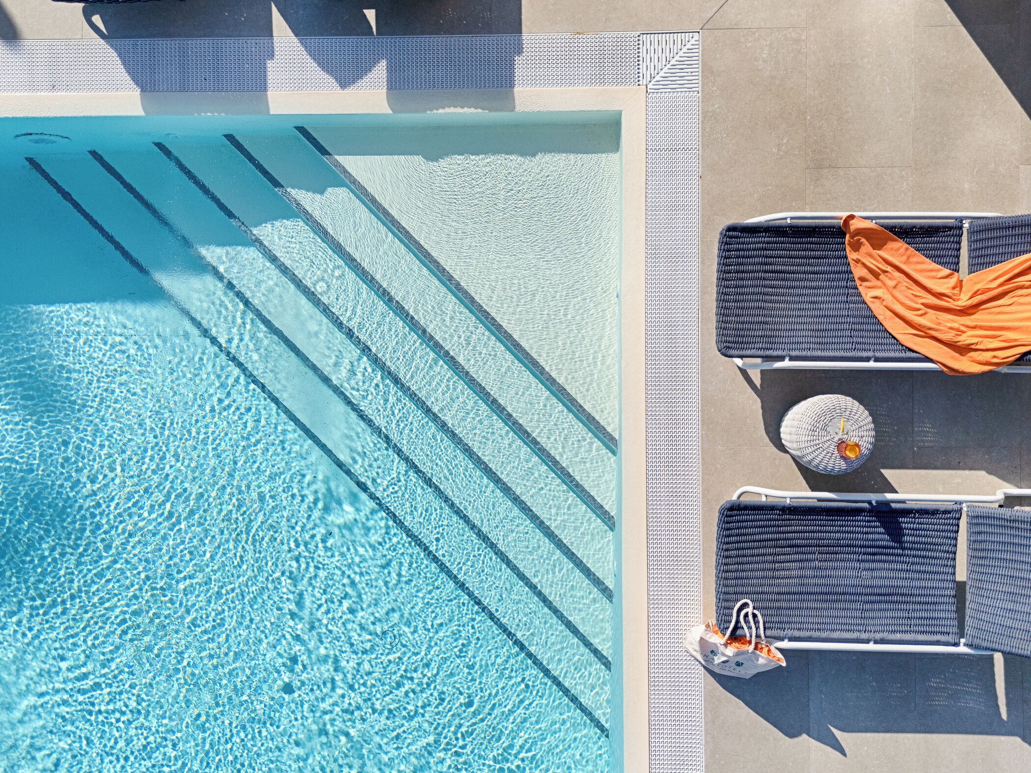 An aerial view of a swimming pool with clear water, adjacent lounge chairs, and a textured walkway.