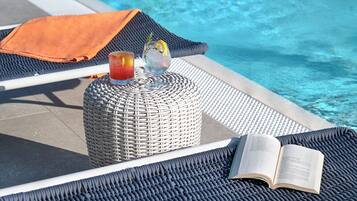 Area relax a bordo piscina con un poggiapiedi in rattan, un drink e un libro aperto.