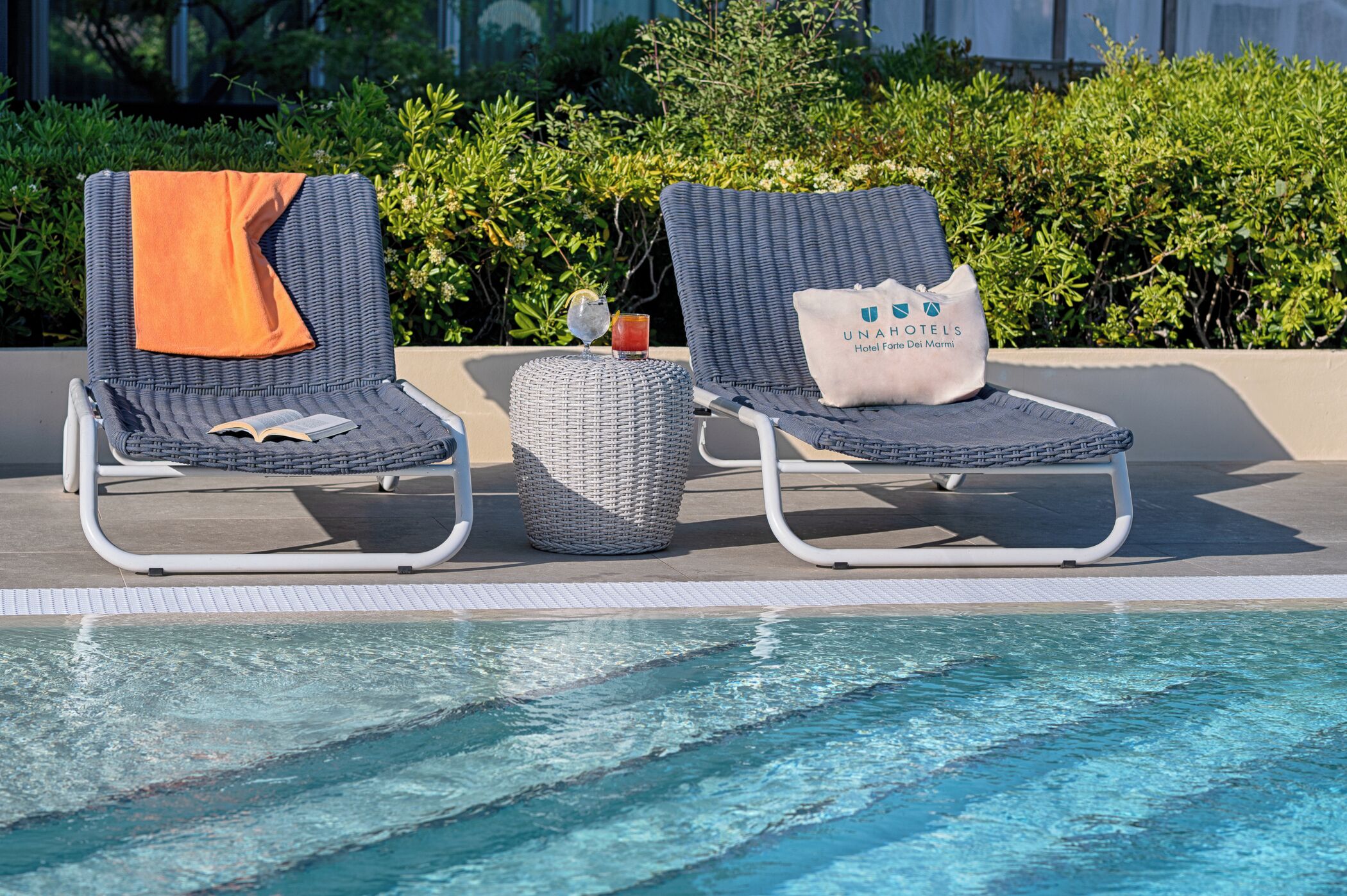 Two lounge chairs by a pool with a book and a glass of drink on one, and a pillow with a logo on the other.