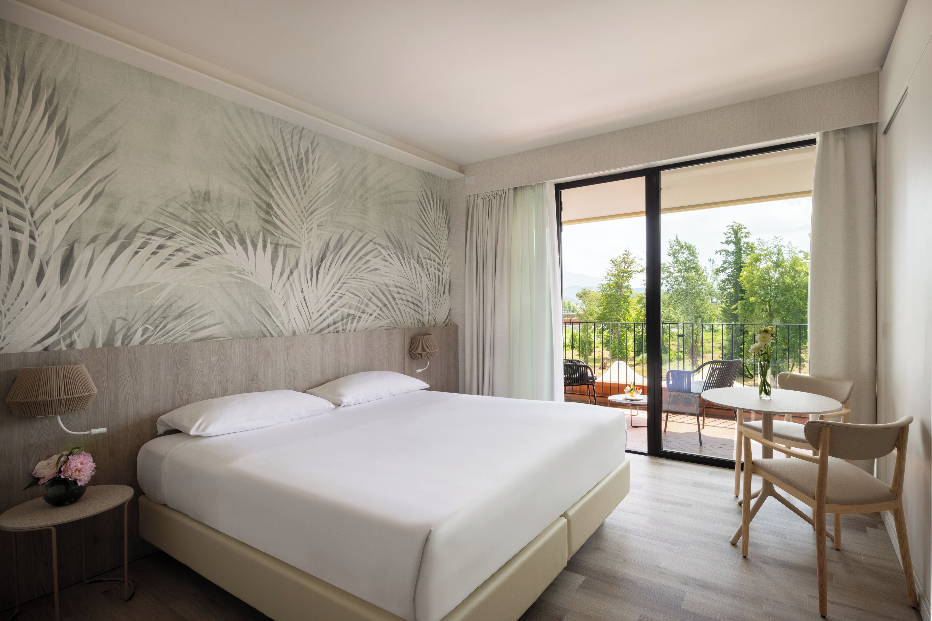 A hotel room with a large bed, a small side table with a vase of flowers, a wall-mounted lamp, and a balcony with a table and chairs.
