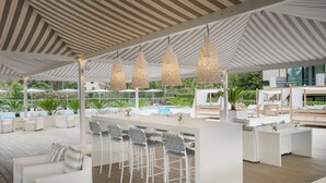 Uma área de bar moderna ao ar livre, com móveis brancos, luminárias pendentes e uma piscina ao fundo.