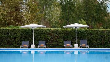 Uma área à beira da piscina com quatro espreguiçadeiras e dois guarda-sóis, cercada por um sebe e árvores.