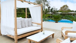 Uma cama de dossel de madeira com quatro colunas, roupa de cama branca, uma mesa de mármore e um ambiente à beira da piscina com guarda-sóis e vegetação.