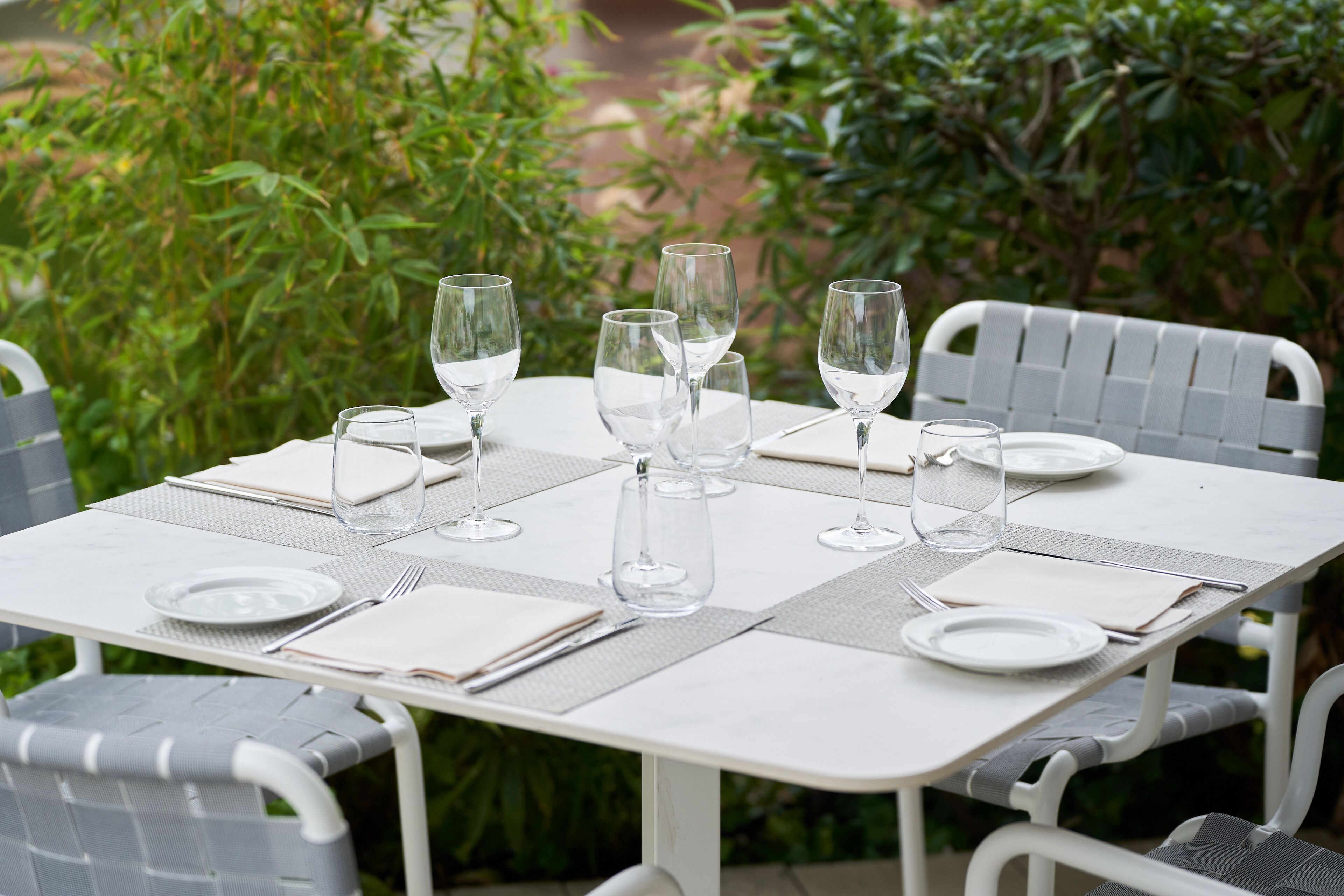 A table set outdoors with clear glassware, plates, and cutlery, surrounded by greenery.
