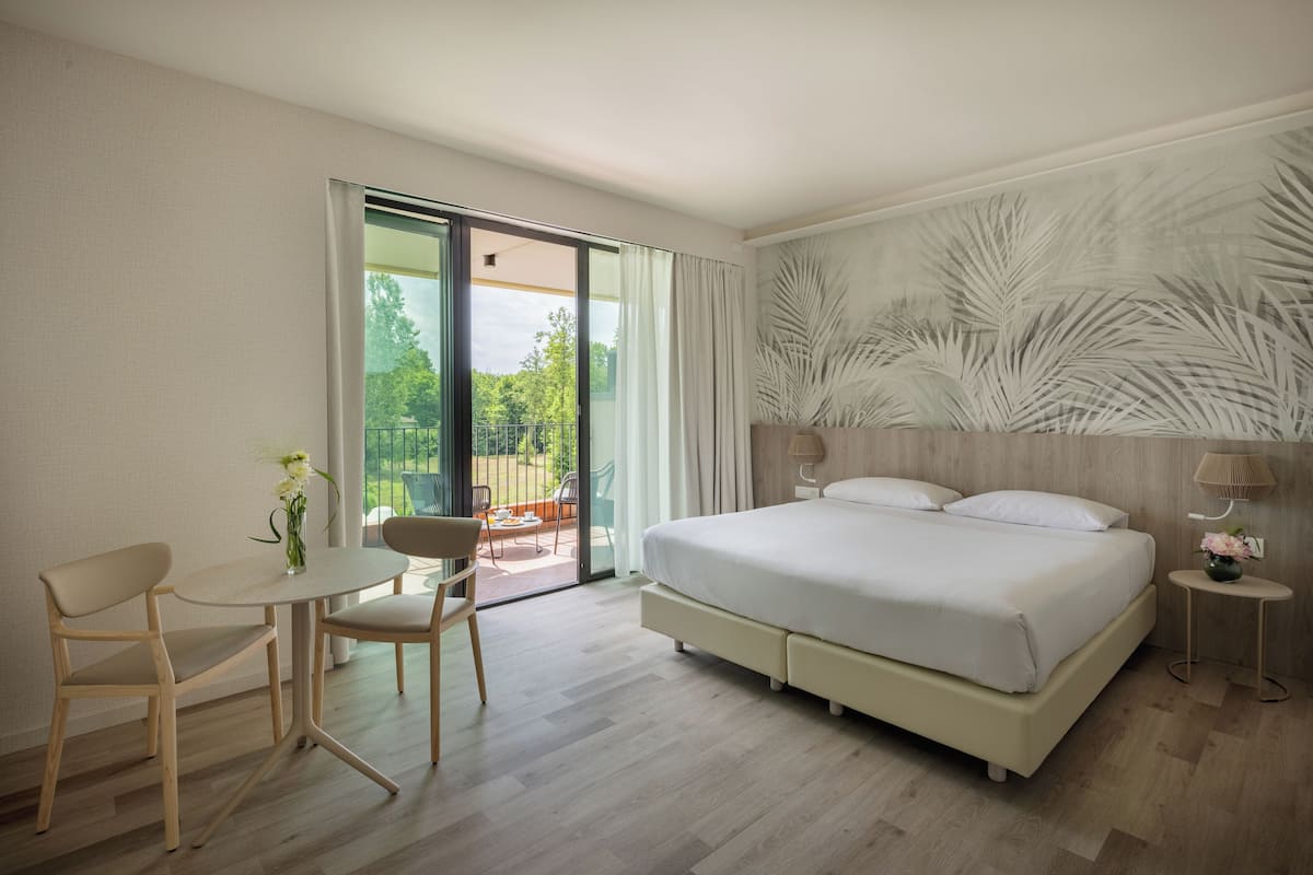 A modern hotel room with a large bed, a small table with chairs, a balcony with a view of greenery, and a wall with a leaf pattern.
