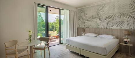 A modern hotel room with a large bed, a small table with chairs, a balcony with a view of greenery, and a wall with a leaf pattern.