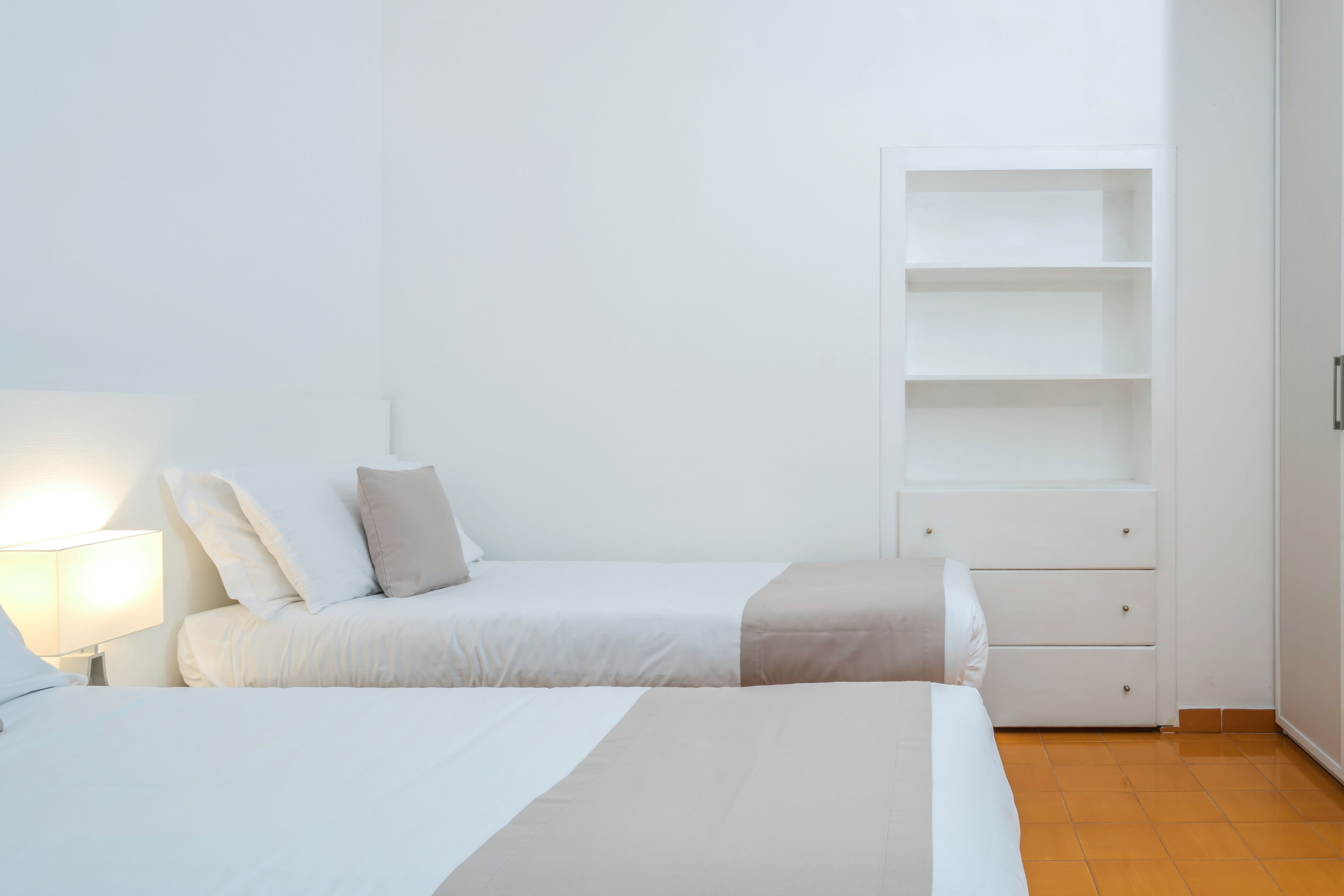 A minimalist bedroom with two beds, a bedside table with a lamp, and a white wardrobe with drawers.
