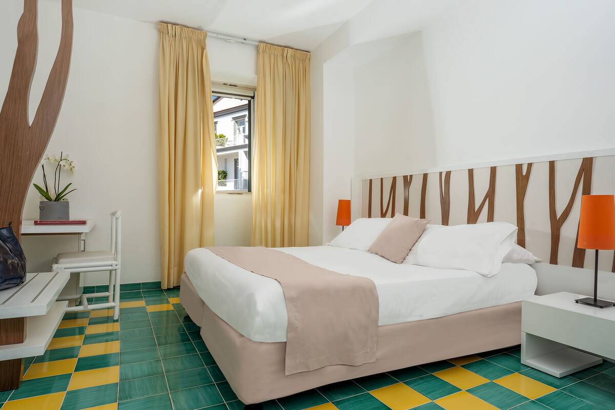 A hotel room with a bed, a desk, a chair, a potted plant, and a window with curtains.
