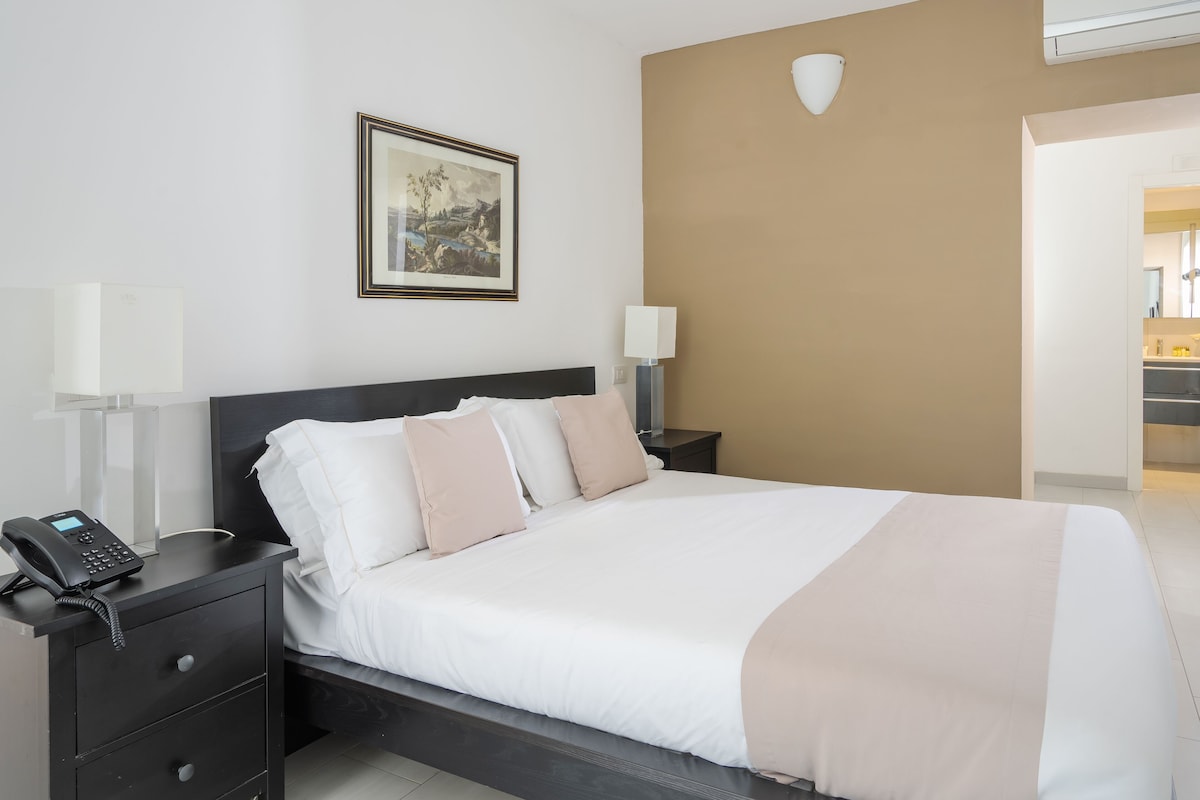 A neatly made bed with a white and beige bedspread, a black nightstand with a telephone, and a framed picture on the wall.
