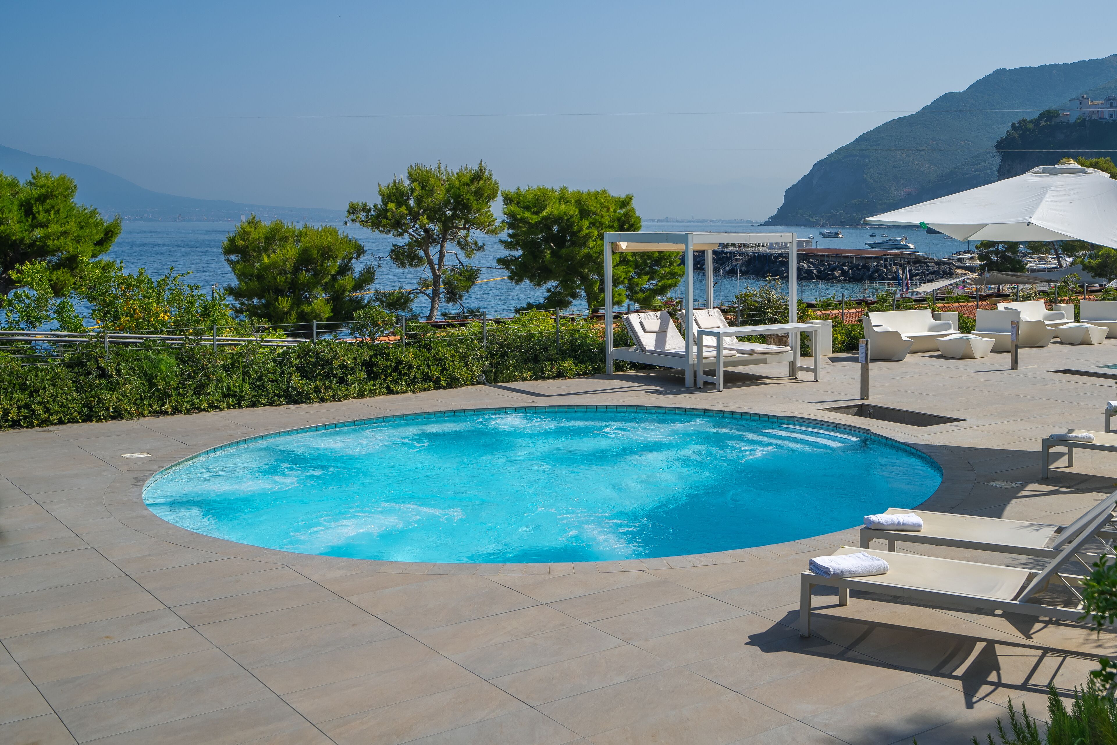 A swimming pool with lounge chairs and umbrellas, surrounded by greenery and a view of the sea.