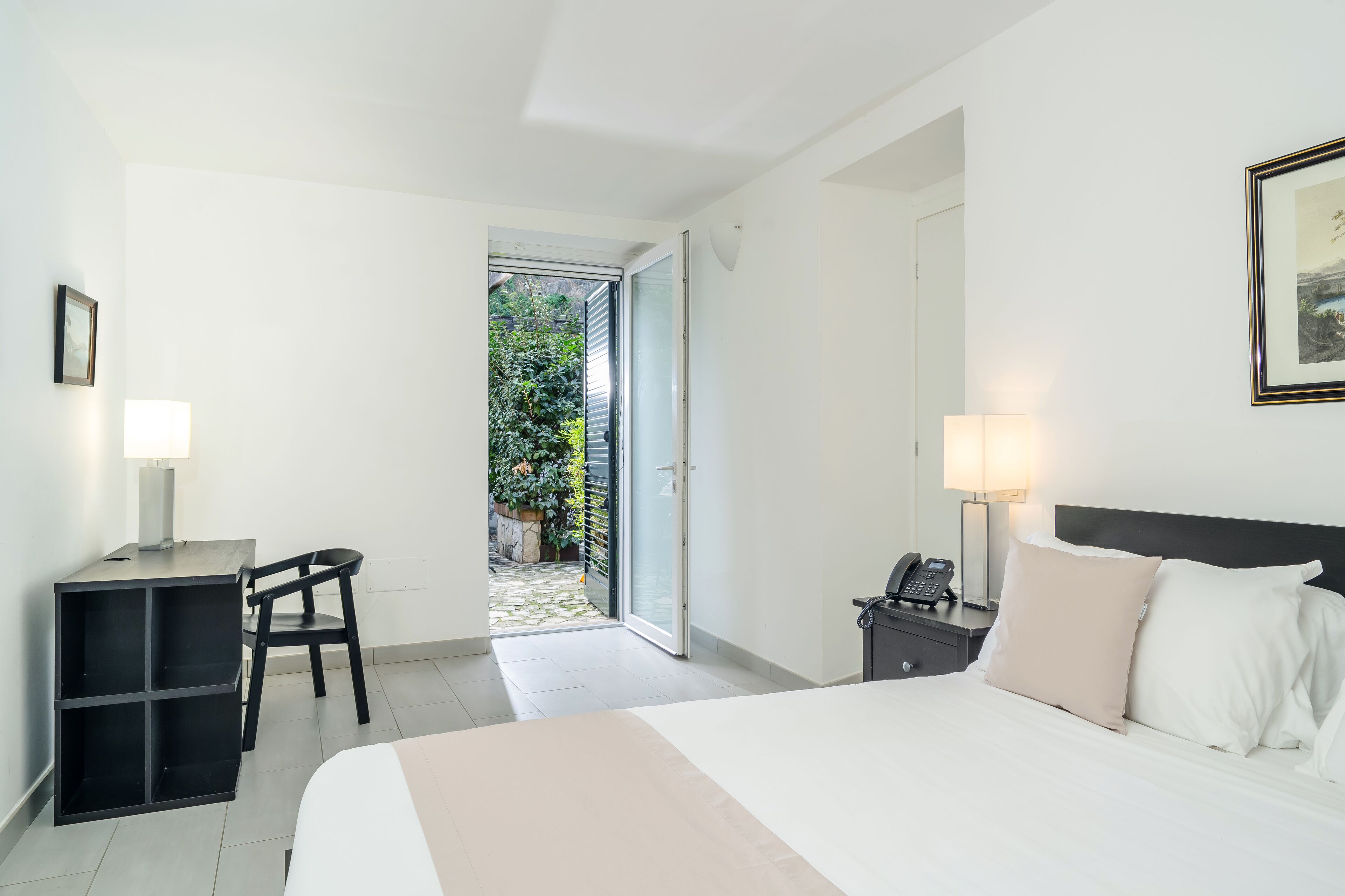 Ein Hotelzimmer mit Bett, Schreibtisch, Stuhl und Blick auf einen Garten durch eine offene Tür.