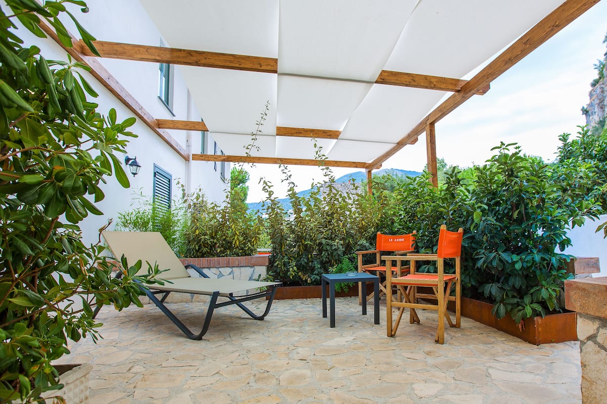 An outdoor patio with a wooden pergola, stone flooring, and various seating arrangements including a lounge chair and dining chairs.